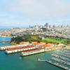 Aerial view of San Francisco waterfront with marinas, piers, and green parks in the foreground, and the city skyline featuring tall buildings in the background under a partly cloudy sky.