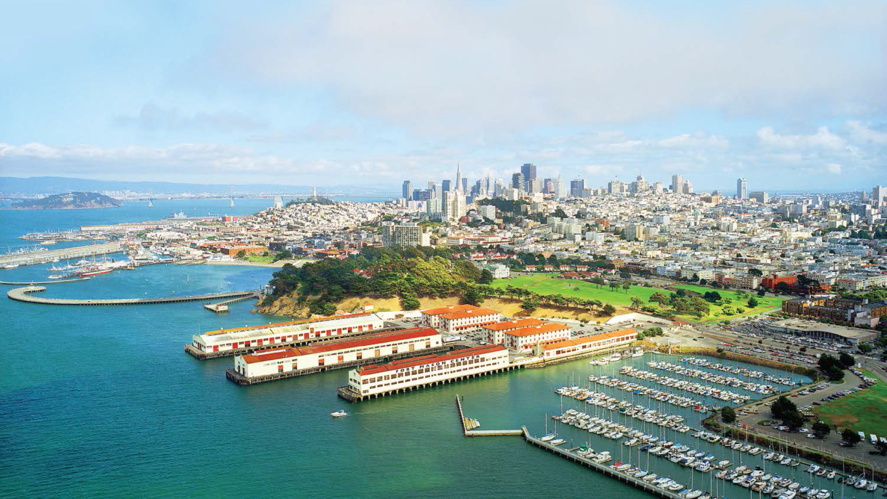 Aerial view of San Francisco waterfront with marinas, piers, and green parks in the foreground, and the city skyline featuring tall buildings in the background under a partly cloudy sky.