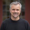 A middle-aged man with short gray hair and a beard smiles at the camera. He is wearing a black sweater and standing in front of a blurred background with muted colors.