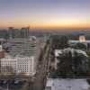 Aerial view of downtown Sacramento at sunset, showing city buildings, tree-lined streets, parked cars, and the illuminated California State Capitol building on the right. The sky has a warm orange and blue gradient.