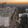 Aerial view of downtown Sacramento at sunset, showing city buildings, tree-lined streets, parked cars, and the illuminated California State Capitol building on the right. The sky has a warm orange and blue gradient.