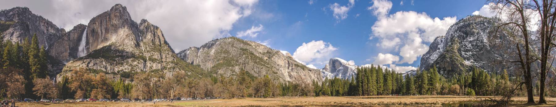 Panoramic view of Yosemite Valley showing granite cliffs, forested areas, open meadows, and bright clouds in a blue sky. Cars are visible parked along the valley floor at the base of the mountains.