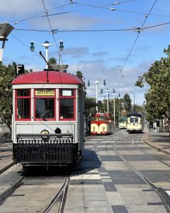 A red and white vintage streetcar travels along curved tracks on a sunny day, with two more streetcars visible further down the track. Trees and lampposts line the tracks under a partly cloudy sky.