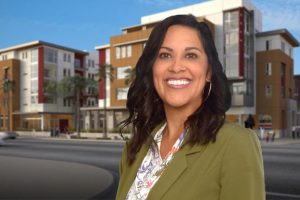 A woman with dark wavy hair, wearing a green blazer and floral blouse, smiles in front of a modern apartment building with palm trees and a clear blue sky.