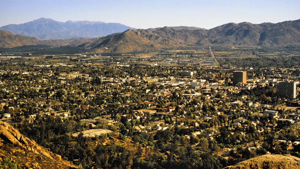 A panoramic view of a city with scattered buildings and houses, surrounded by green trees and mountains in the background under a clear blue sky.