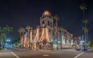 A large, ornate building with a domed tower and arched windows is lit up at night, adorned with string lights and palm trees lining the street. The scene appears calm with little to no traffic or pedestrians.