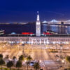 A night view of San Francisco’s Ferry Building with bright lights, palm trees, and the Bay Bridge in the background reflecting on the water. The scene is vibrant and illuminated against a dark sky.