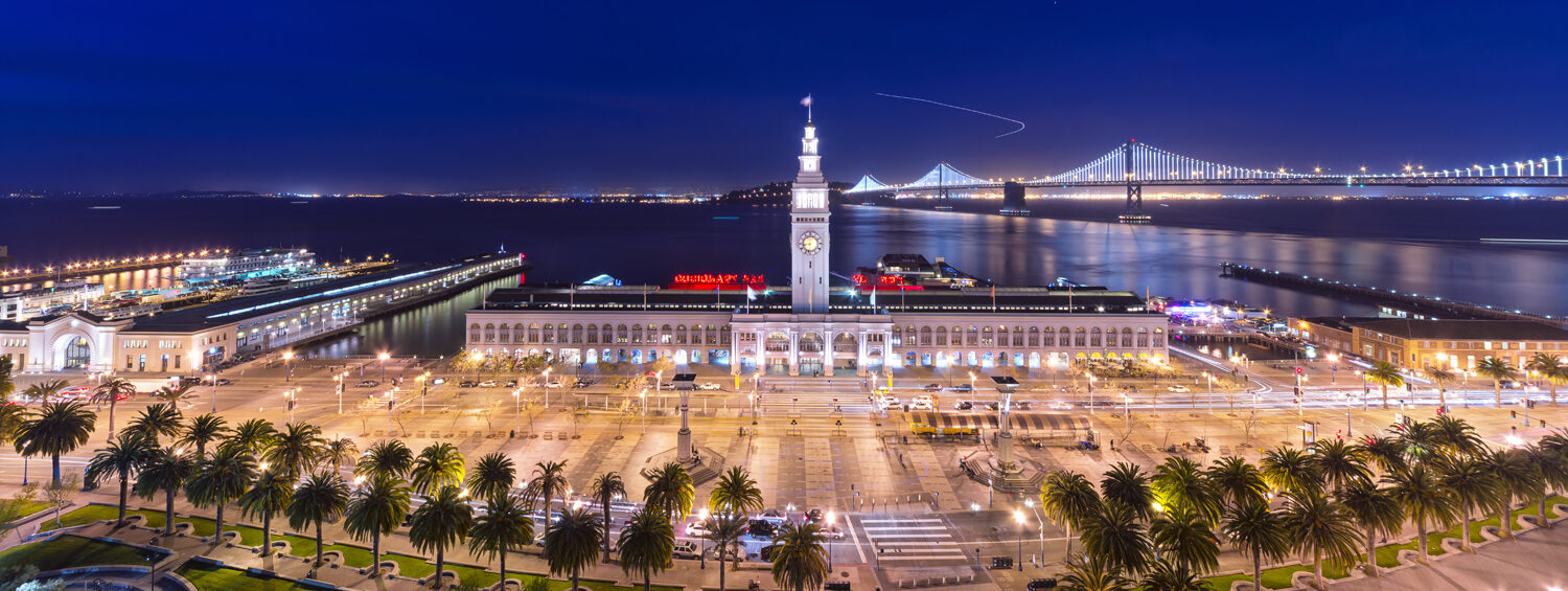 A night view of San Francisco’s Ferry Building with bright lights, palm trees, and the Bay Bridge in the background reflecting on the water. The scene is vibrant and illuminated against a dark sky.