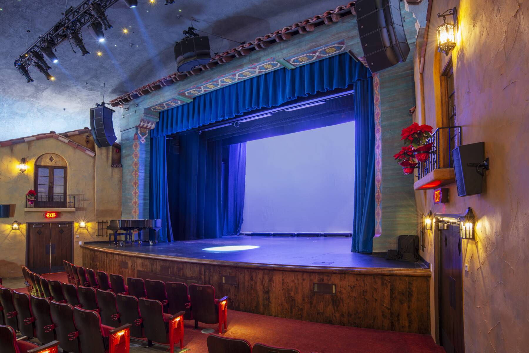 An empty theater with red velvet seats faces a stage framed by blue curtains and decorative walls, lit by wall sconces and ceiling lights, creating a warm and inviting atmosphere.