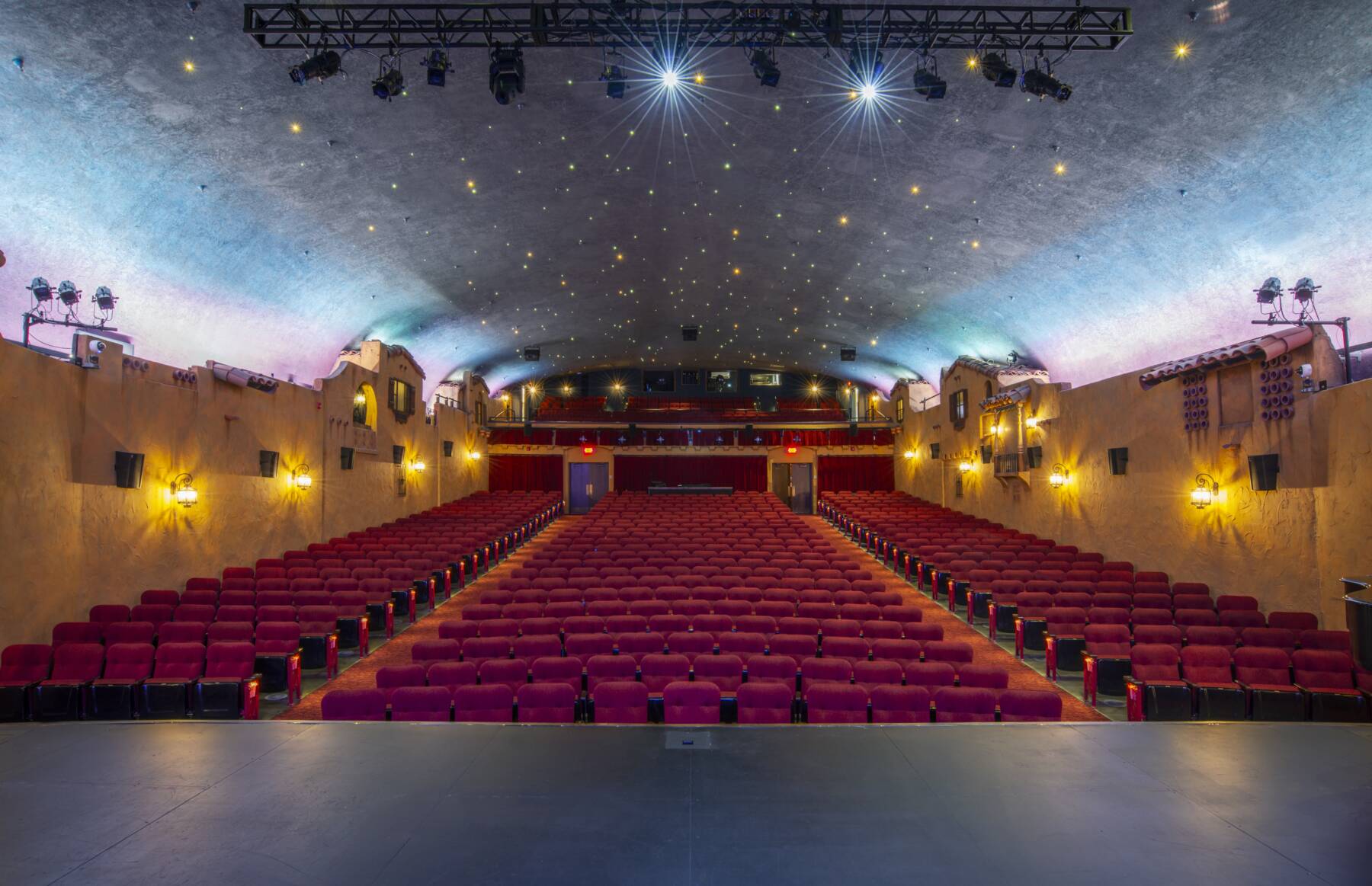 View from a theater stage facing rows of empty red seats, with warm yellow wall lights, decorative walls, and a curved, star-lit ceiling. The auditorium is well-lit and appears ready for an audience.