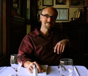 A man wearing glasses and a maroon shirt sits at a restaurant table with two empty wine glasses, a salt shaker, and a folded napkin, smiling slightly in a warmly lit, cozy setting.