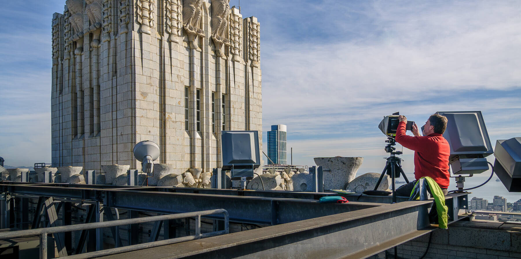 A person in a red jacket uses a large camera on a rooftop near a stone building decorated with eagle sculptures under a blue sky. City buildings are visible in the background.