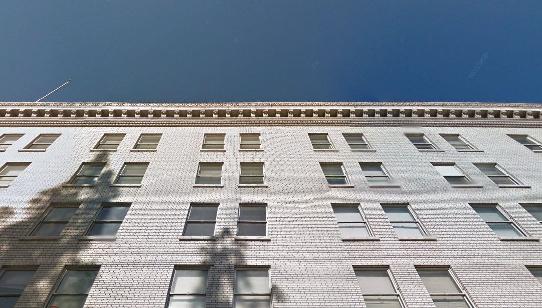 A white brick apartment building with multiple rows of windows, viewed from a low angle against a clear blue sky. Shadows of trees are visible on the building’s facade.