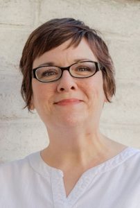 A woman with short brown hair and glasses, wearing a white shirt, smiles softly while standing in front of a light-colored brick wall.
