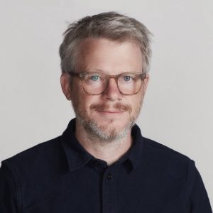 A man with light brown hair and a short beard, wearing glasses and a dark collared shirt, smiles slightly while looking at the camera against a plain light background.
