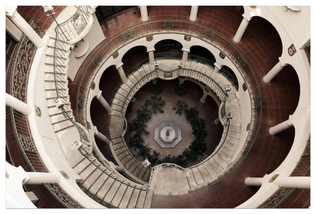 A view from above of a circular, multi-level courtyard with ornate railings, white columns, red tile floors, and a central fountain surrounded by greenery. Spiral staircases connect the different levels.