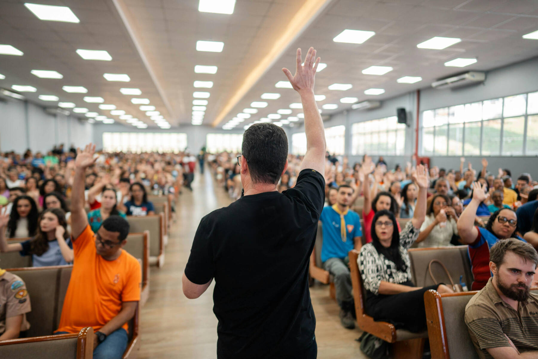 A man stands at the front of a large room, facing an audience with raised hands. The audience is seated in rows, engaged and attentive, inside a brightly lit space with large windows.