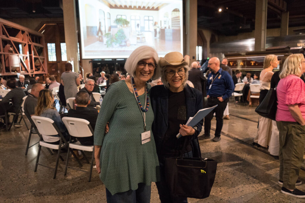 Two smiling older women stand together in a busy indoor event space, surrounded by people sitting and standing. A large screen with an image is projected in the background. Both women wear name tags.