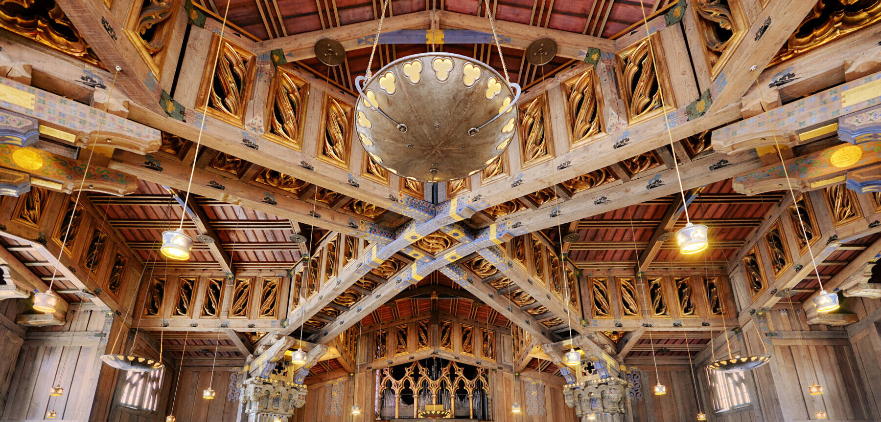 An ornate church interior features wooden beams, gold accents, columns, hanging lights, and rows of wooden pews. A few people are seated, facing the altar at the front. The ceiling and walls display intricate architectural details.