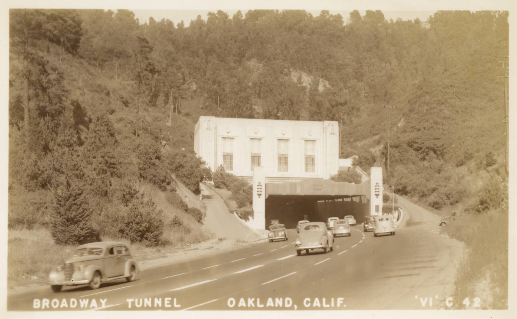 Vintage photo of cars driving into the Broadway Tunnel in Oakland, California, surrounded by trees and hills; large white tunnel entrance is prominent in the center.