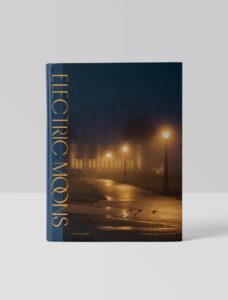A book titled Electric Moons stands upright. The cover shows a foggy night scene with glowing street lamps lining a wet pathway, leading to a building adorned with lights.