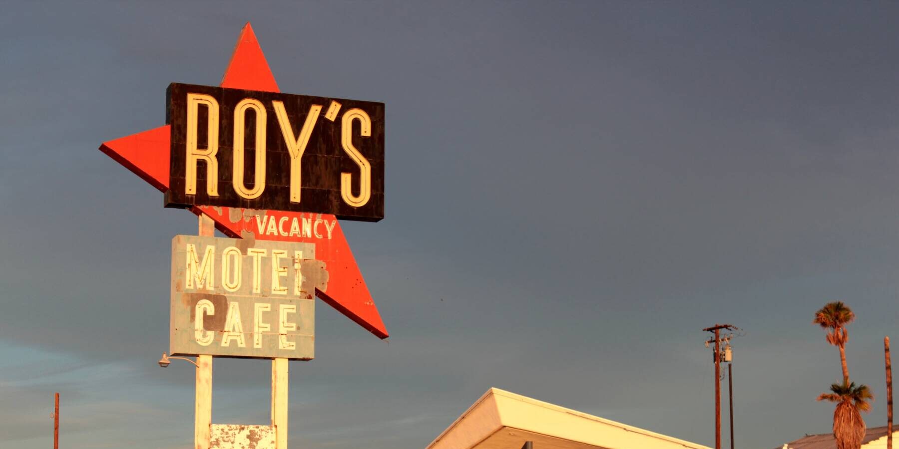 A vintage roadside sign reading Roys Motel Café stands beside a mid-century style building under a clear sky, with palm trees and desert plants in the background.