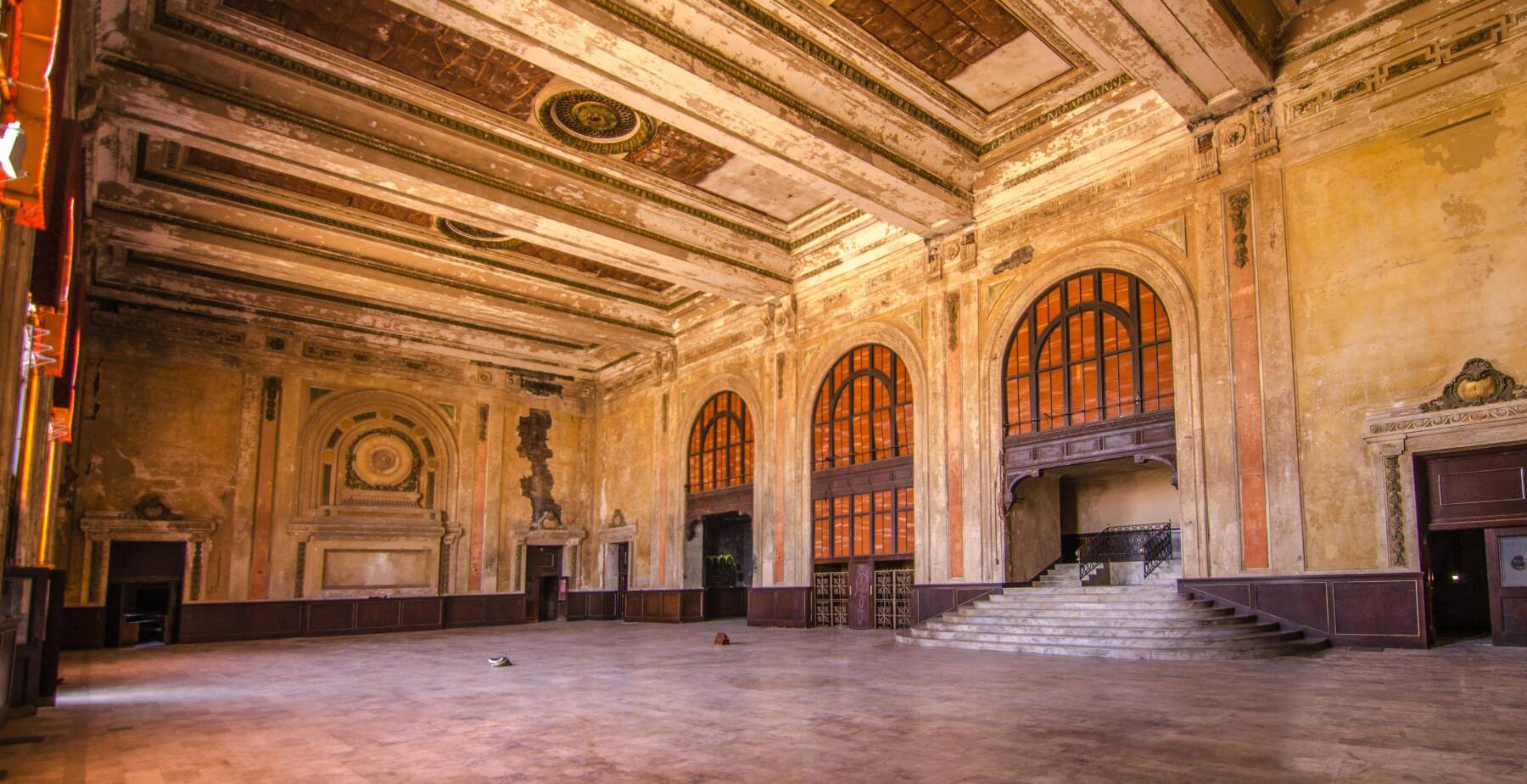 A large, empty, ornate hall with tall arched windows, faded yellow walls, a decorated high ceiling, and a grand staircase. Light streams in, illuminating the worn interior and scattered objects on the floor.