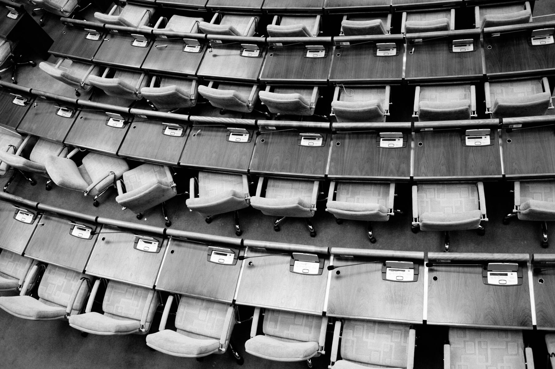 Black and white photo of several rows of empty seats and desks in an auditorium or legislative chamber, viewed from above. Each desk has a microphone and a built-in device.