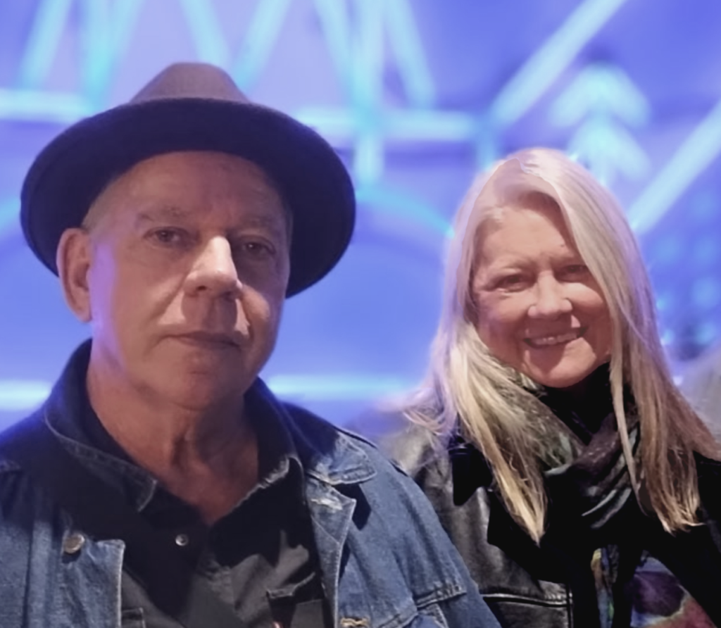 A man in a denim jacket and black hat stands next to a smiling woman with long blonde hair, both in front of a blue-lit geometric background.