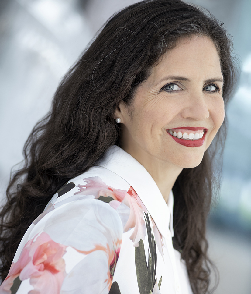 A woman with long dark hair, wearing a white blouse with a floral pattern, smiles at the camera against a blurred, light-colored background.