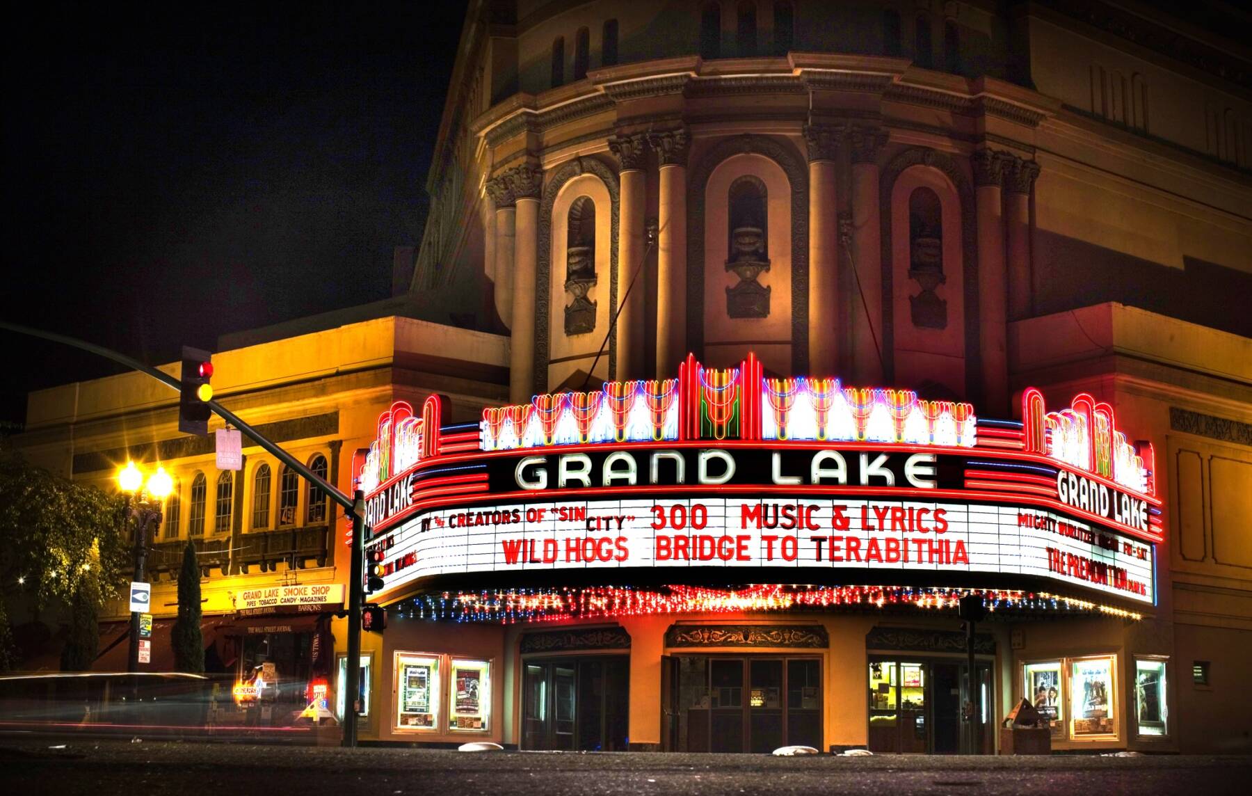A brightly lit Grand Lake theater marquee at night displays movie titles including 300, Music & Lyrics, Wild Hogs, and Bridge to Terabithia. The historic building has ornate architecture and glowing neon lights.
