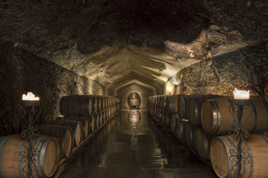 A dimly lit wine cellar tunnel with stacked wooden barrels lining both sides, stone walls and ceiling, and candle sconces illuminating the entrance.