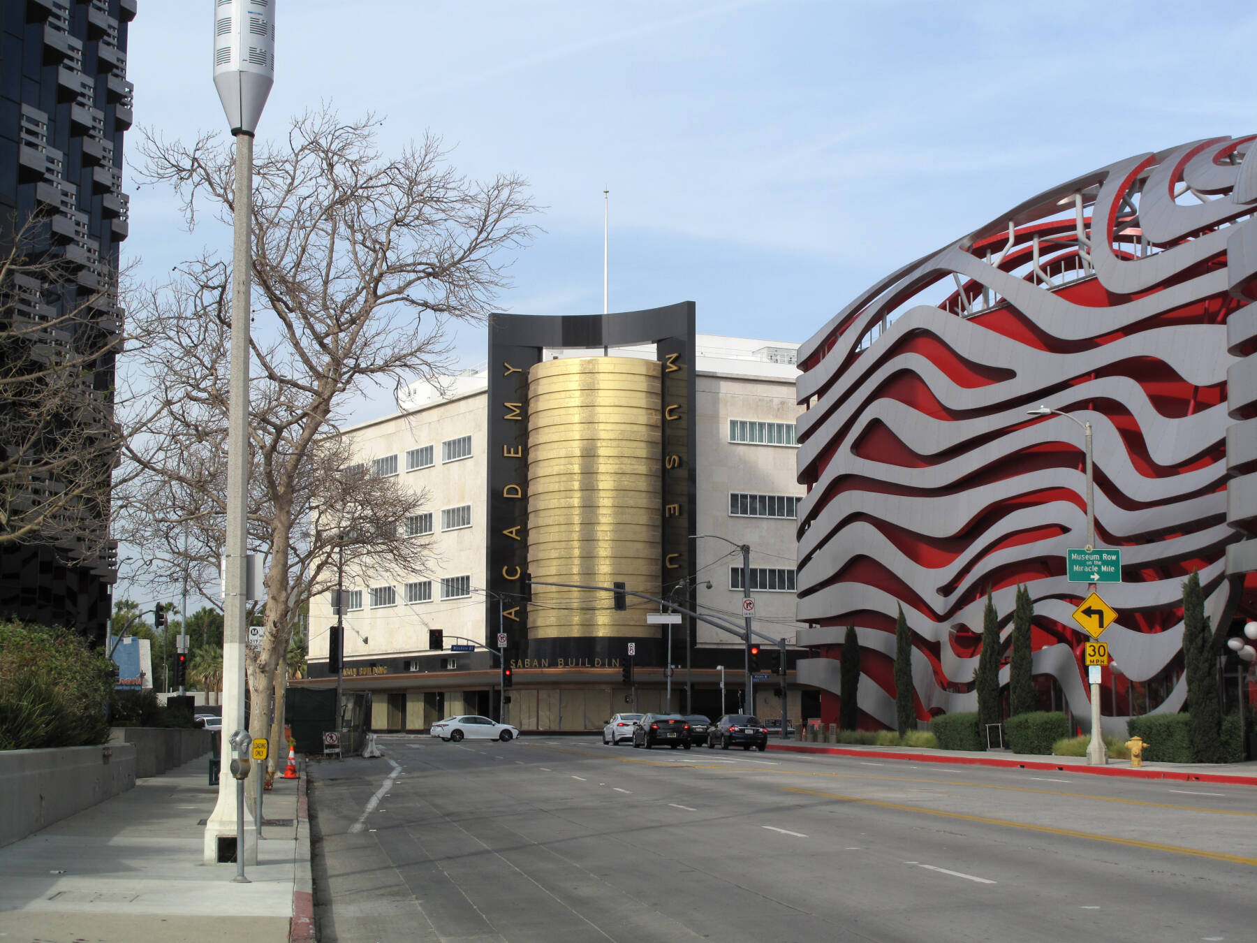 A street view of the Academy Museum of Motion Pictures in Los Angeles, showing its gold entrance and the red-and-white wavy wall design on the adjacent building. Some cars are parked along the road.