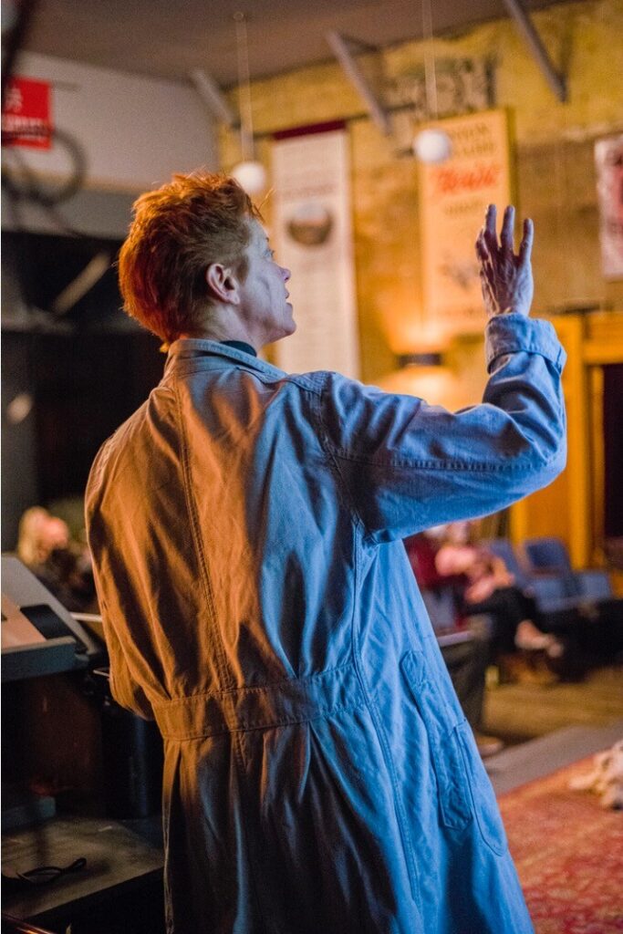 Person with short red hair wearing a light blue coat gestures with one hand while speaking in a room with banners and an audience in the background.