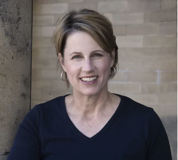 A woman with short, light brown hair and hoop earrings smiles at the camera. She is wearing a black V-neck shirt and is posed in front of a tan brick wall and a stone pillar.
