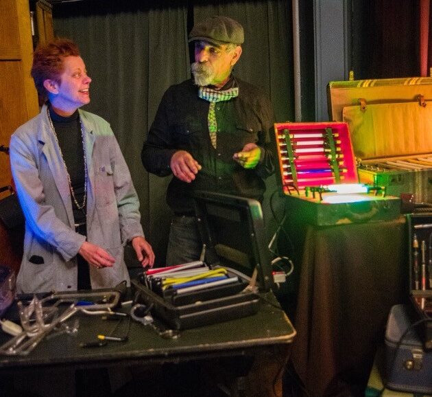 Two people stand by a table with glowing, colorful tubes and equipment cases in a dimly lit room. An EXIT sign is visible above a curtain behind them.