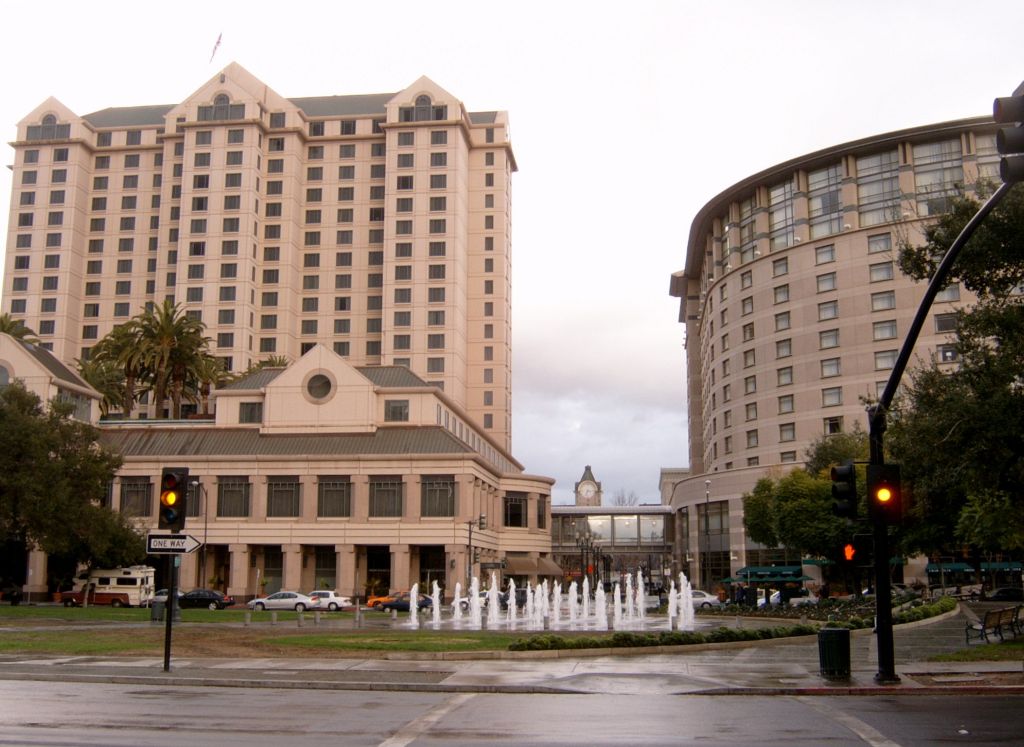 A tall hotel and a curved building stand behind a fountain and traffic lights on a city street. Trees and cars line the area, and the sky is overcast.