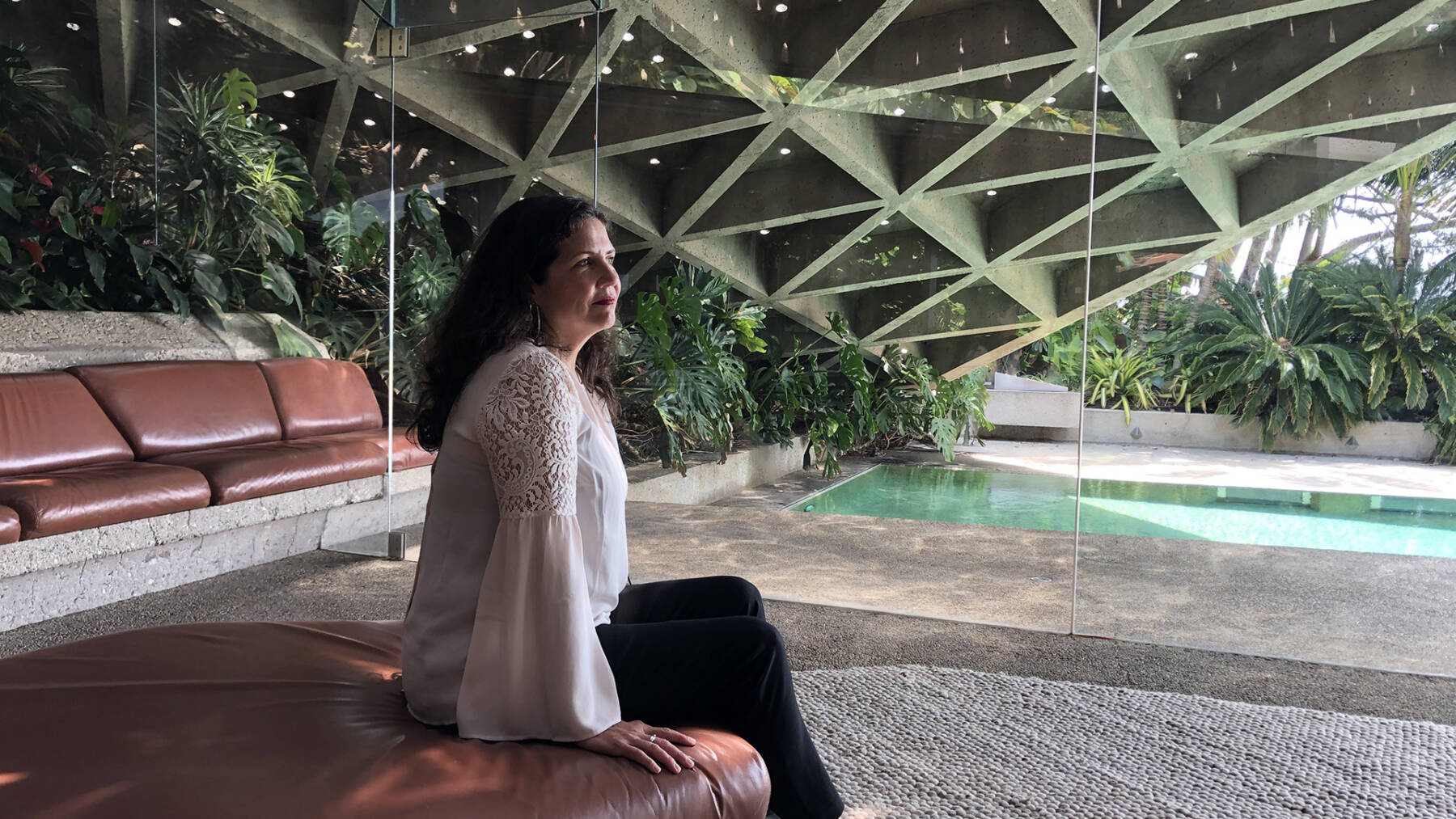 A woman in a white blouse sits on a brown leather couch, gazing out at a lush garden and pool through large glass windows in a modern room with angular ceiling beams.