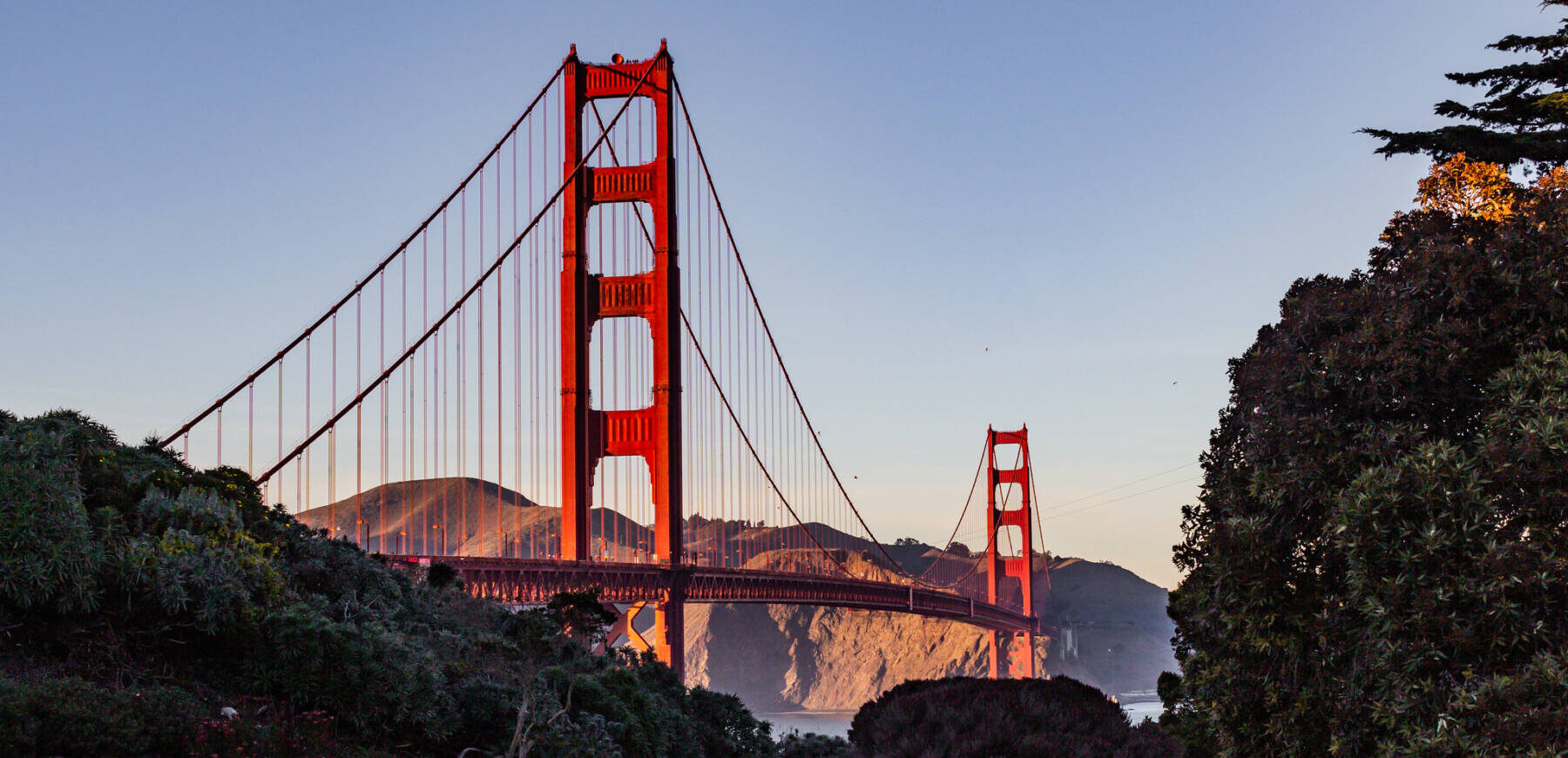The Golden Gate Bridge in San Francisco is shown at sunset, surrounded by greenery, with hills and a clear sky in the background.