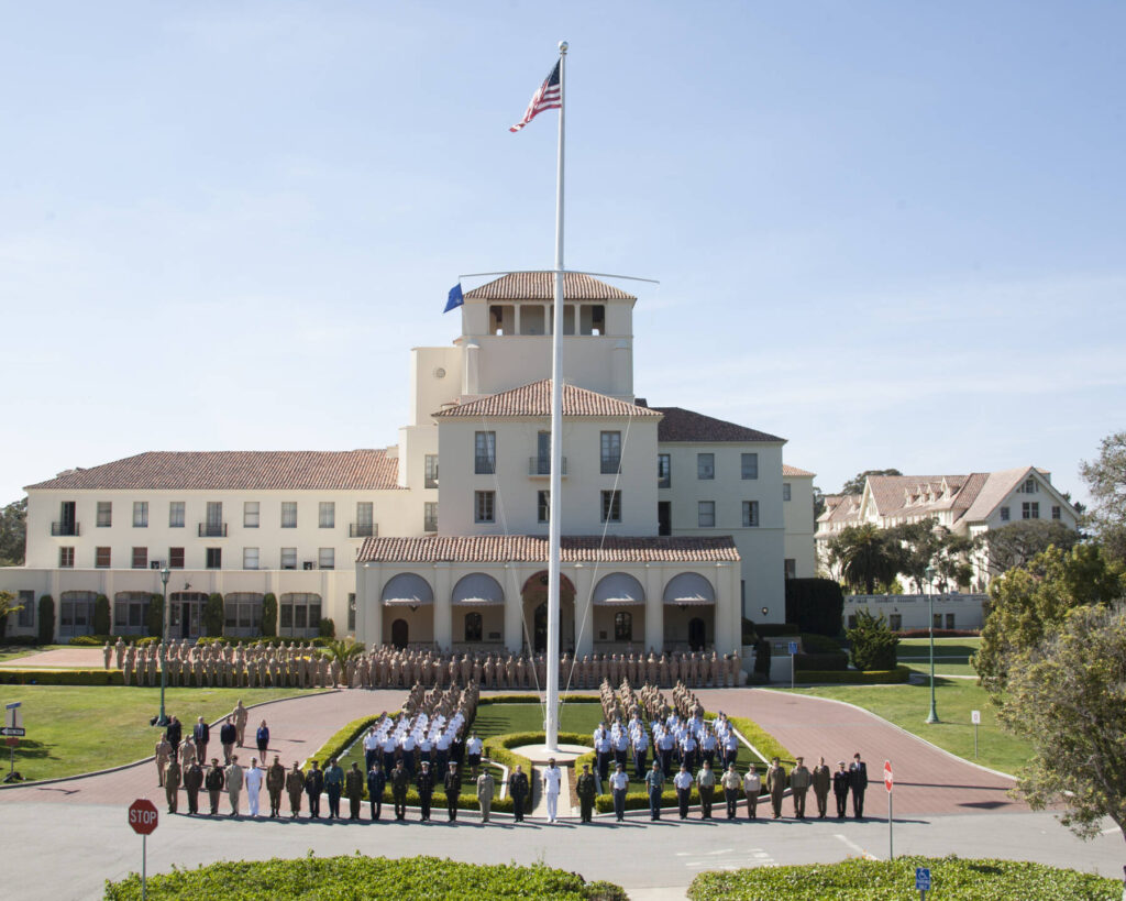 A large group of uniformed cadets stands in formation around a flagpole in front of a white, multi-story building with a red-tiled roof, under a clear blue sky.