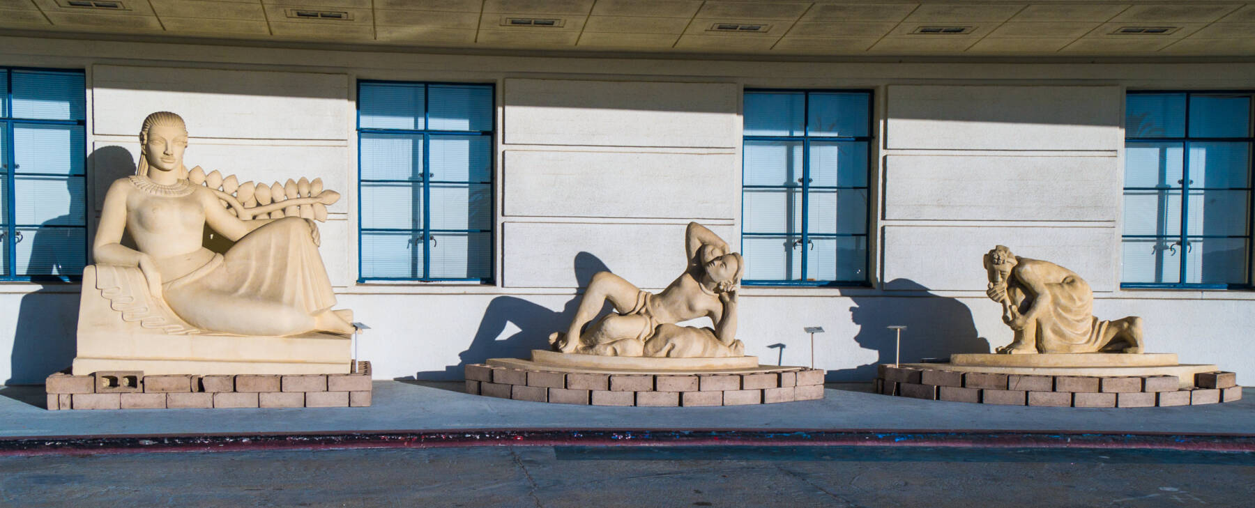 Three large tan sculptures of reclining and kneeling human figures displayed on stone platforms in front of a building with blue-framed windows.