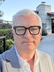 A man with gray hair and black glasses, wearing a light-colored blazer and white shirt, stands outside near a building and greenery on a sunny day.