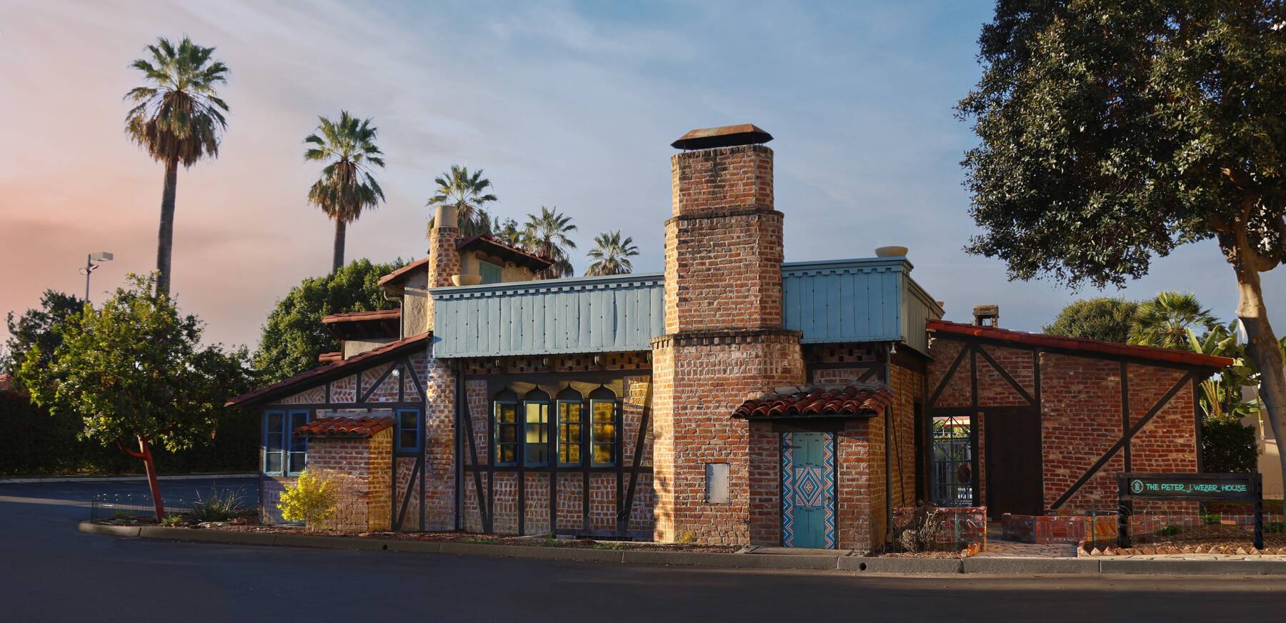 A rustic brick building with a tall chimney, blue-trimmed windows, and tile details, surrounded by palm trees and greenery under a colorful sky.