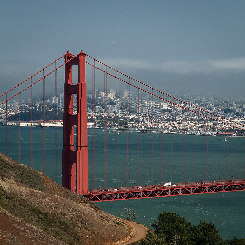 The Golden Gate Bridge spans blue water with the San Francisco city skyline in the background, viewed from a nearby hillside under a clear sky.