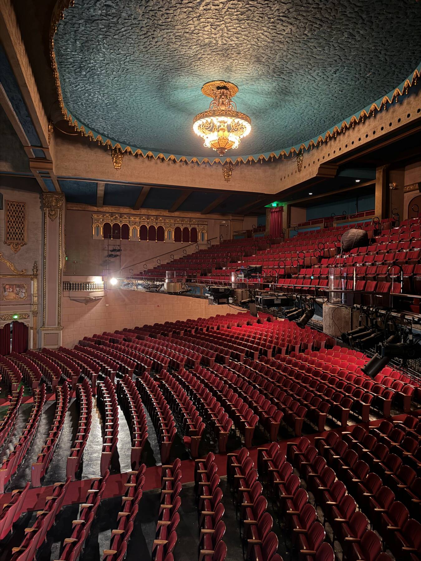 Interior of a historic theater showing an ornate ceiling with a chandelier above rows of empty red seats leading toward a stage.