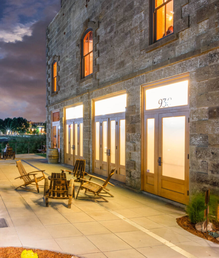 A stone building with a patio featuring wooden chairs and tables overlooking a river at dusk.