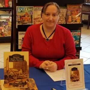 A woman in a red sweater sits at a table with copies of her book, Historic Movie Theatres of West Virginia, and a sign announcing her author event.